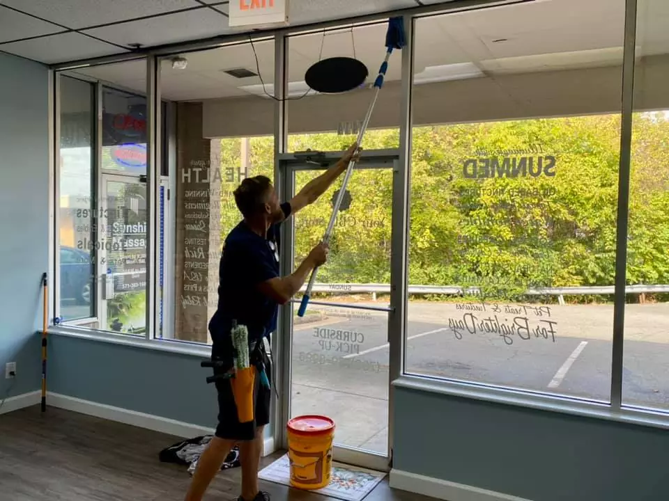 A worker is cleaning the large glass windows of a storefront with a long-handled squeegee inside a building, with the exterior view showing trees and a parking lot.