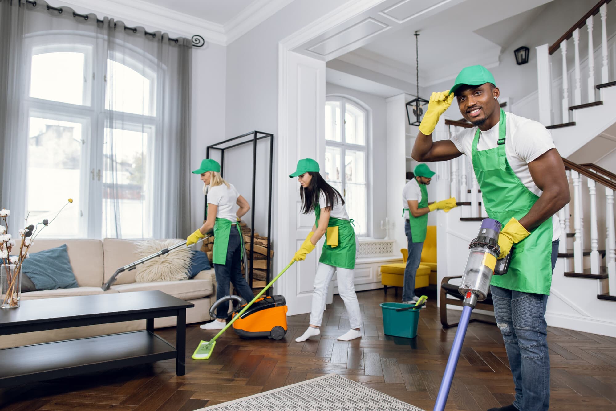 A team of four cleaning professionals in green aprons and hats is engaged in various cleaning tasks throughout a well-lit living room, utilizing equipment like a vacuum and mops.